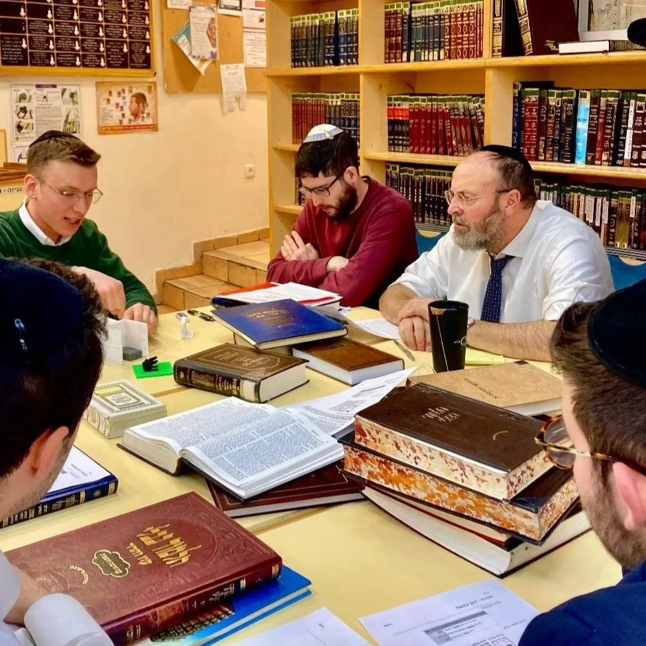 Group of men studying Jewish texts around a table filled with books and papers in a library.