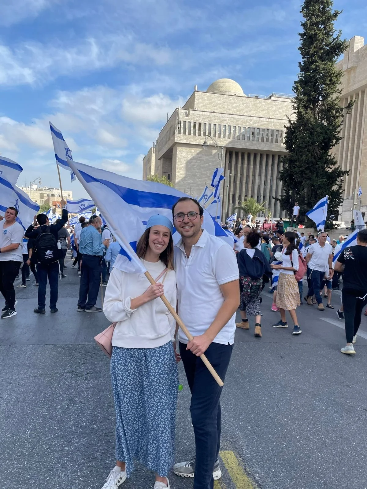 Two smiling people holding Israeli flags at a gathering in an urban area. The woman is wearing a white hoodie, blue patterned skirt, and white sneakers, and the man is wearing a white polo shirt and dark pants. Behind them are many other people, some also holding flags, in front of a large, official-looking building with a dome on top and a tall pine tree to the right.