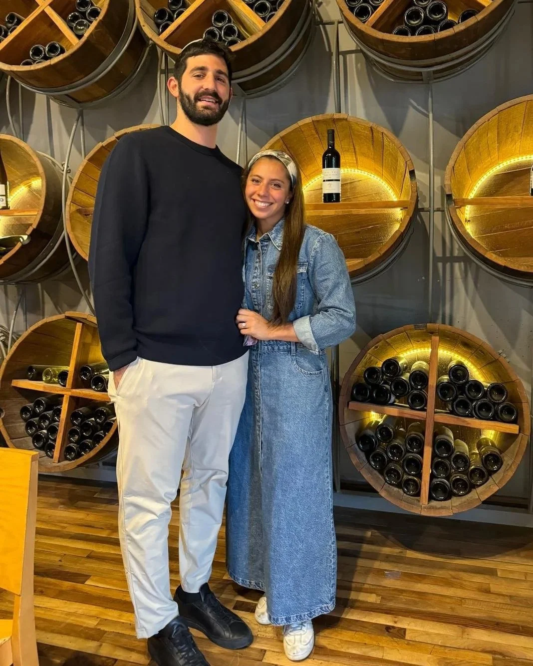 A man and woman stand together, smiling, in front of a wall of wooden wine barrels filled with wine bottles in a winery.
