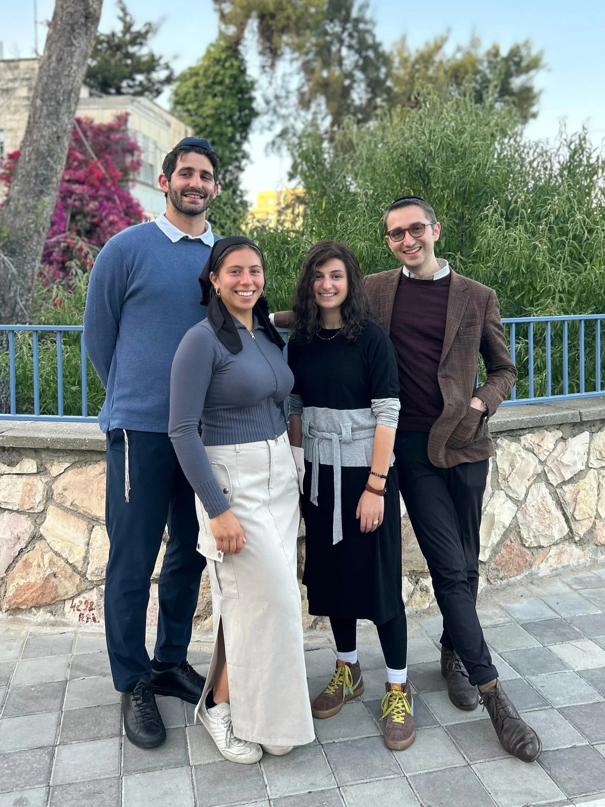 Four people standing together outdoors, smiling. They are on a paved area with a stone wall and green bushes behind them, along with trees and a building. The group consists of two men and two women, wearing casual and smart-casual clothing.