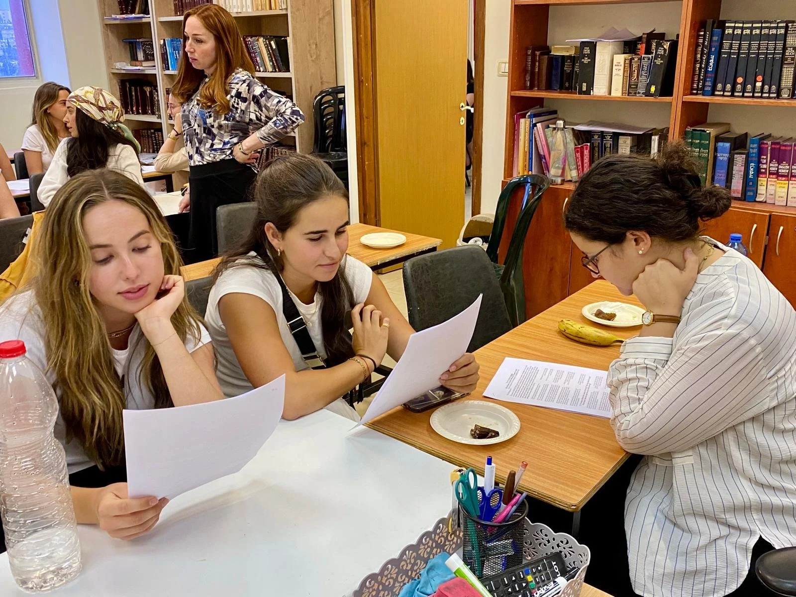 Three women sitting at a table in a library or classroom, looking at papers. One woman is reading, and another is resting her chin on her hand. In the background are bookshelves and other women sitting at tables.