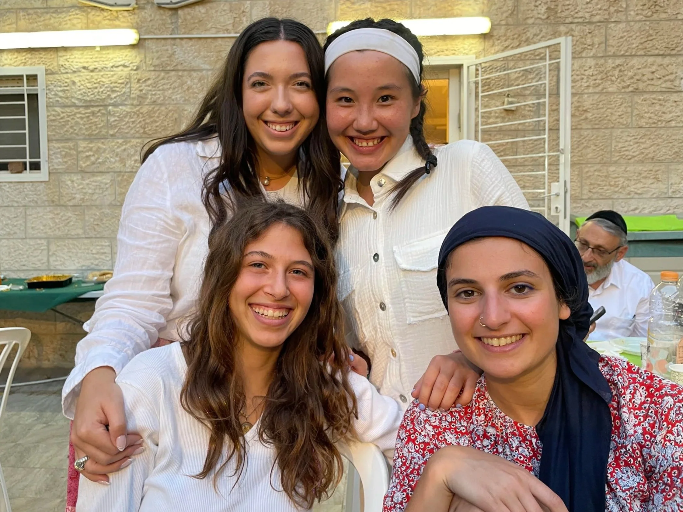Group of smiling women gathered closely outdoors, with a beige stone wall and window in the background, enjoying a social event.