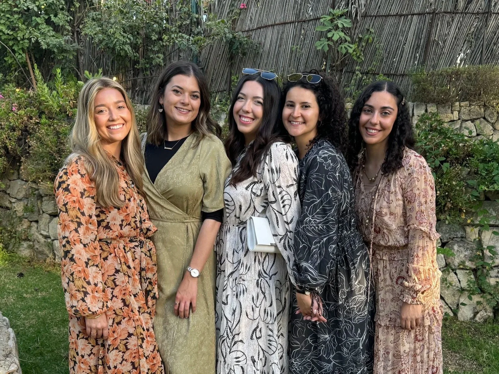Five women standing together outdoors in front of a stone wall and greenery, smiling at the camera. They are wearing dresses with various floral and leafy patterns, and some have sunglasses on their heads.