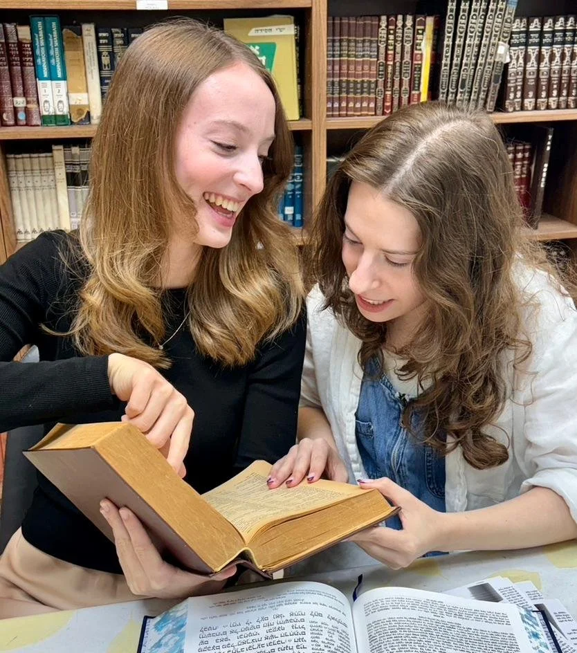 Two young women reading a book together in a library, with shelves of books in the background, smiling and engaged in conversation.