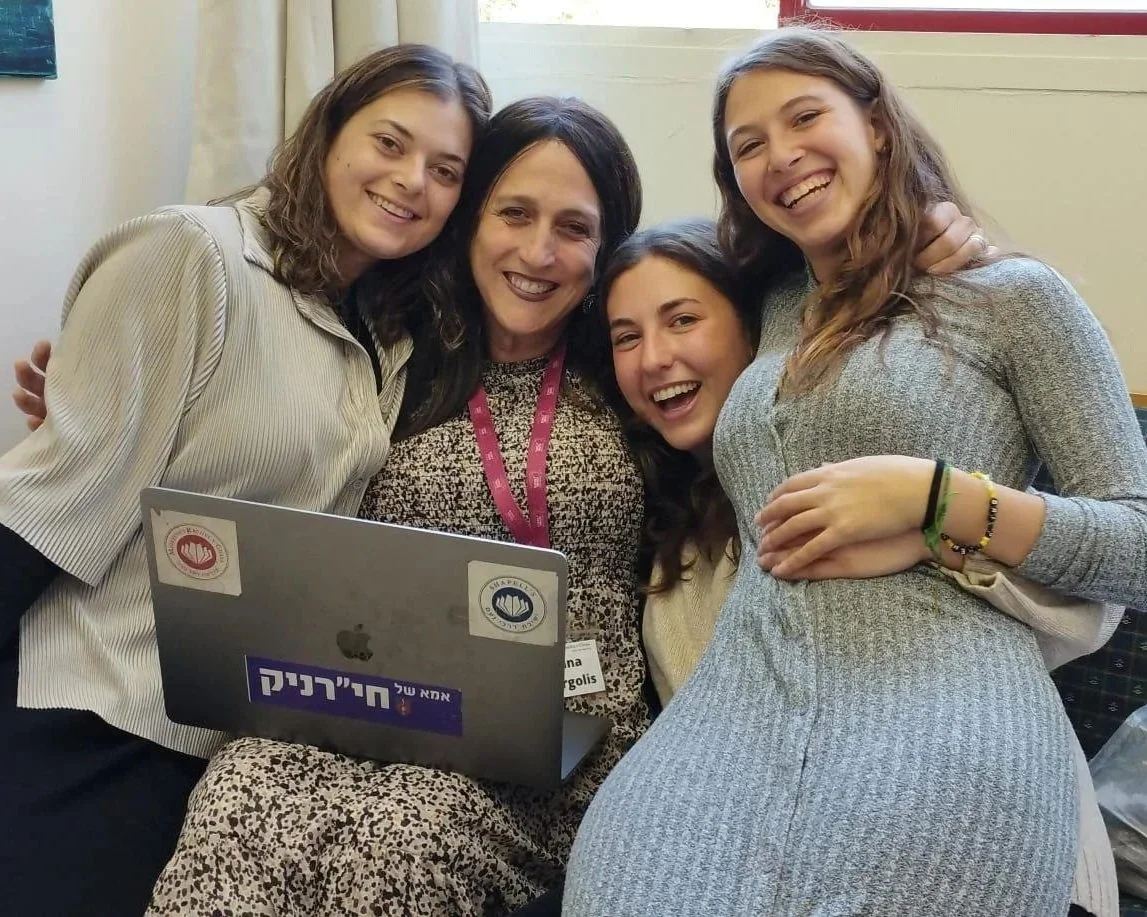 Four women smiling and hugging in front of a laptop at a classroom or office setting.