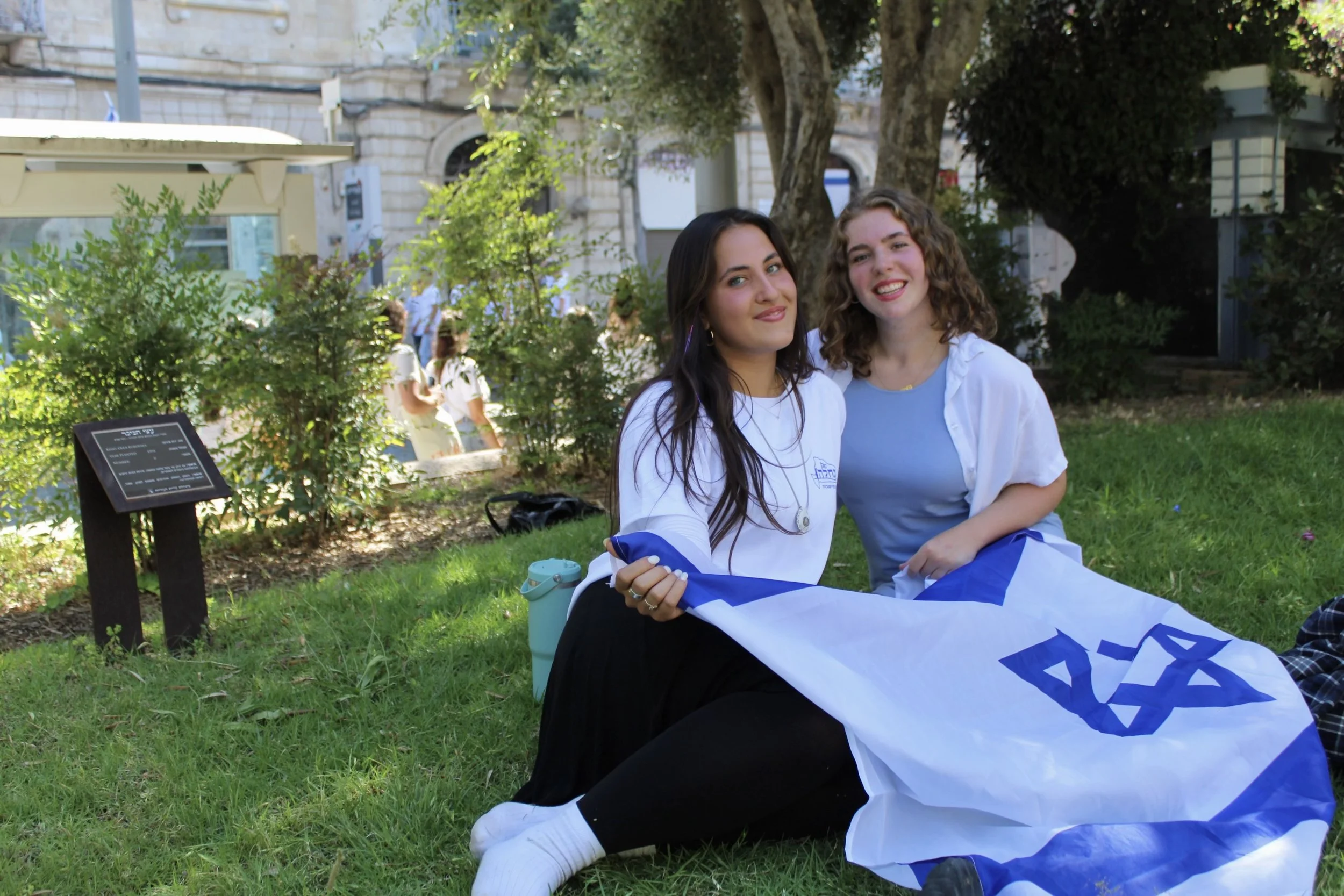 Two young women sitting outdoors on the grass, smiling, with a flag and a black signboard nearby, surrounded by green foliage and trees in an urban setting.