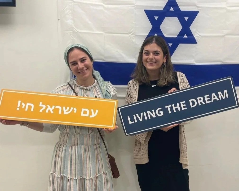 Two women standing in front of an Israeli flag holding signs that say 'Living the Dream' and a sign with Hebrew text.