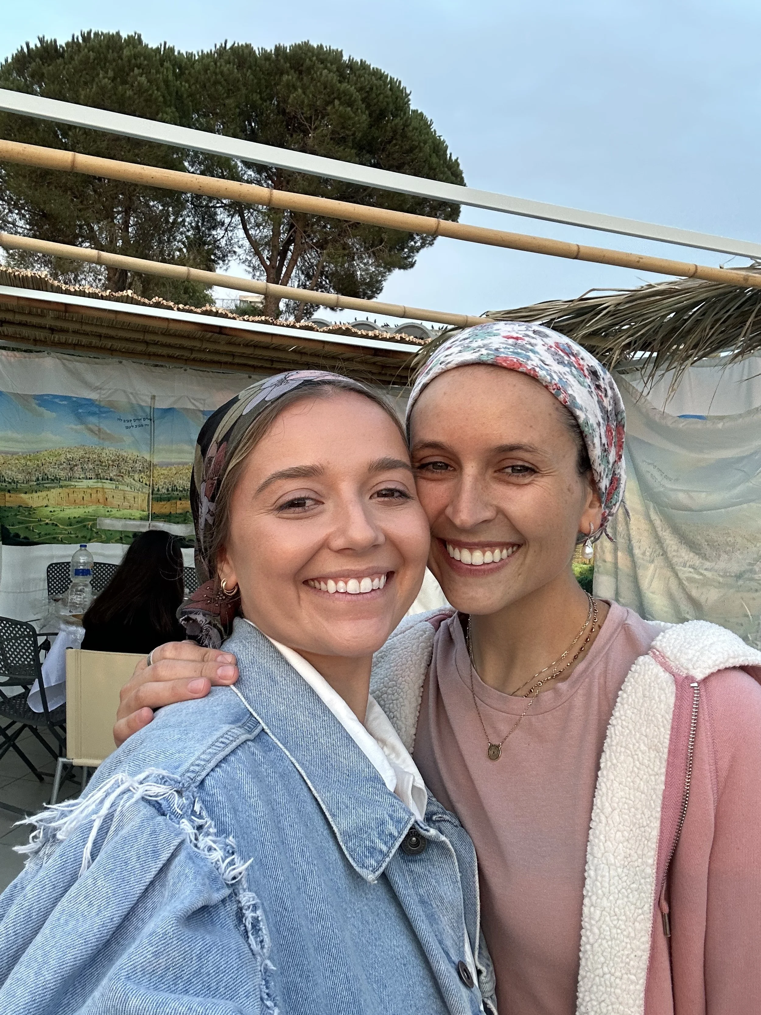 Two smiling women taking a selfie outdoors, with a large tree and a tent or canopy in the background.