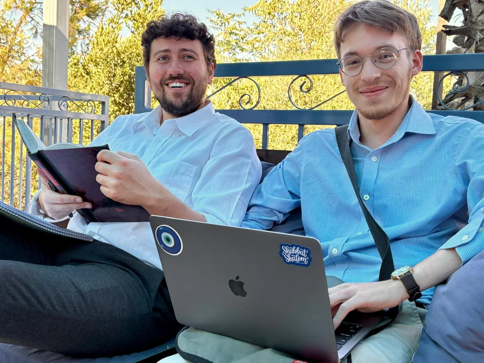 Two young men sitting outdoors on a bench, smiling at the camera, with laptops on their laps. One is holding a book.