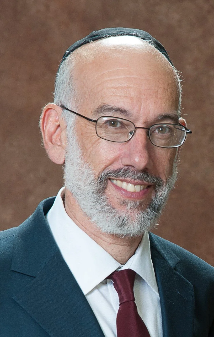 Close-up portrait of a smiling elder man with glasses, a white beard, and a kippah, wearing a suit and tie, against a brown background.
