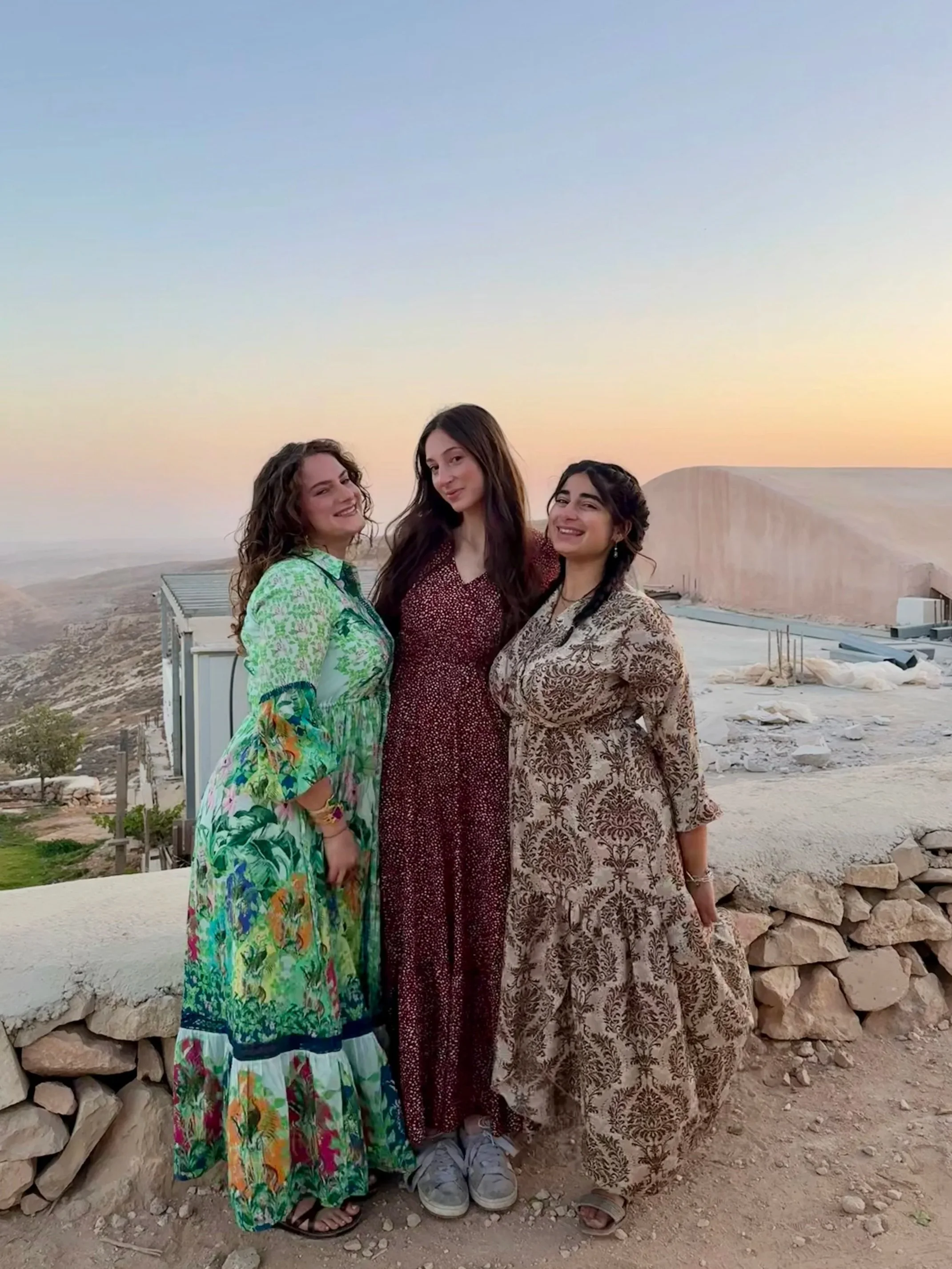 Three women standing together outdoors during sunset, wearing colorful and patterned dresses, with a construction site and rocky landscape in the background.