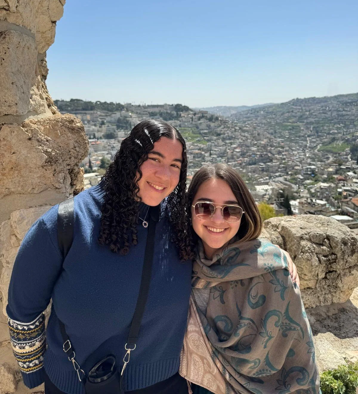 Two smiling women standing close together outdoors, with a cityscape and hillside in the background, near a stone wall.