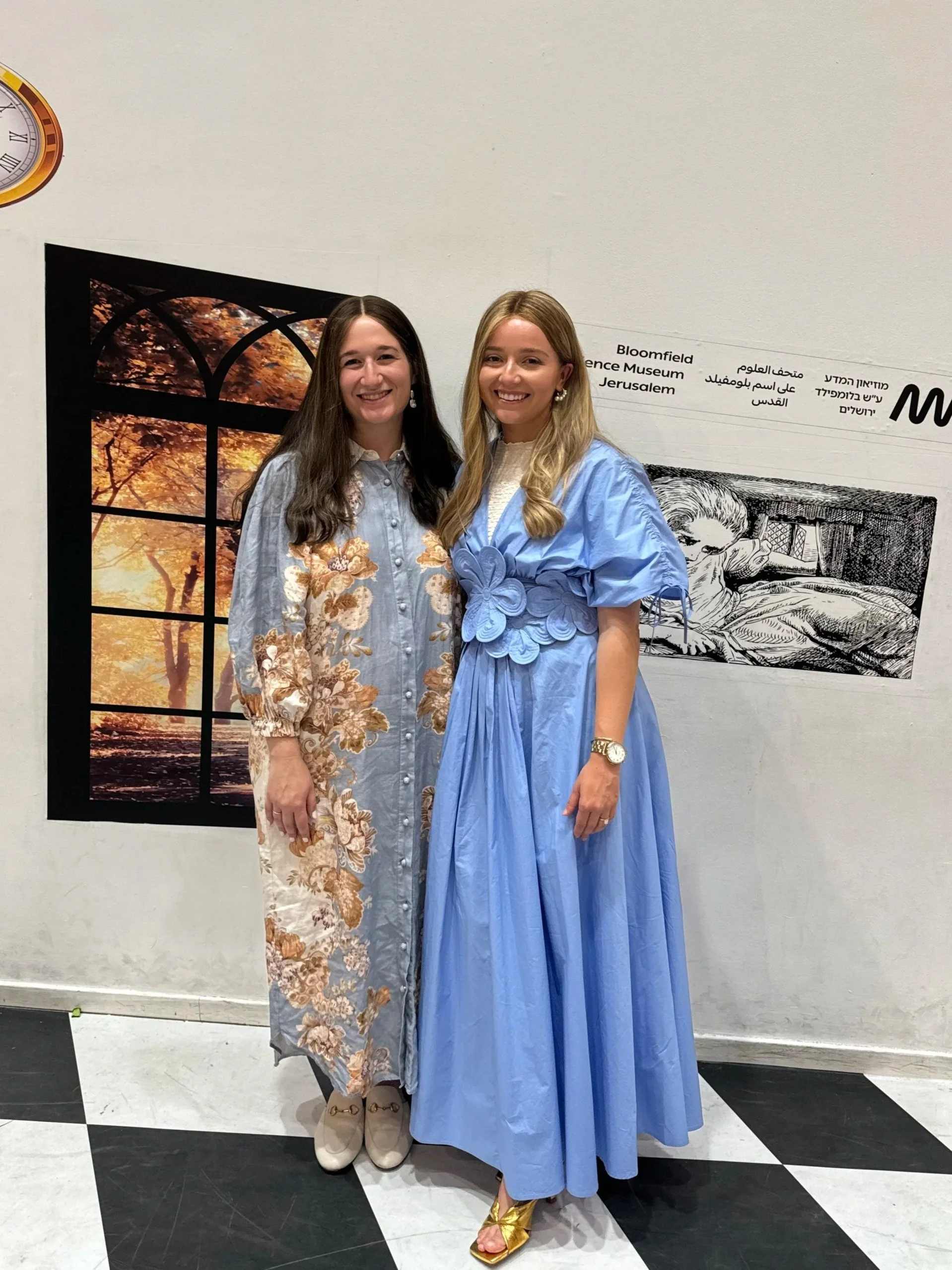 Two women standing next to each other smiling at a museum exhibit. One woman has dark hair and is wearing a long floral dress, and the other woman has blonde hair and is wearing a light blue gown and gold sandals.
