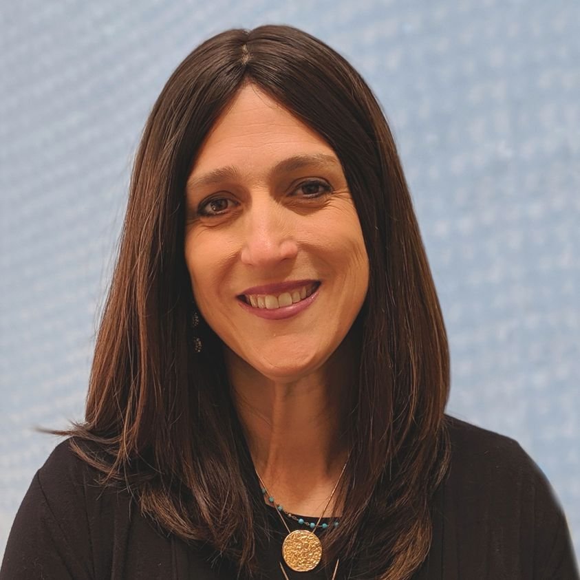 A woman with shoulder-length dark brown hair, smiling, wearing a black top and layered necklaces, standing in front of a light blue textured background.