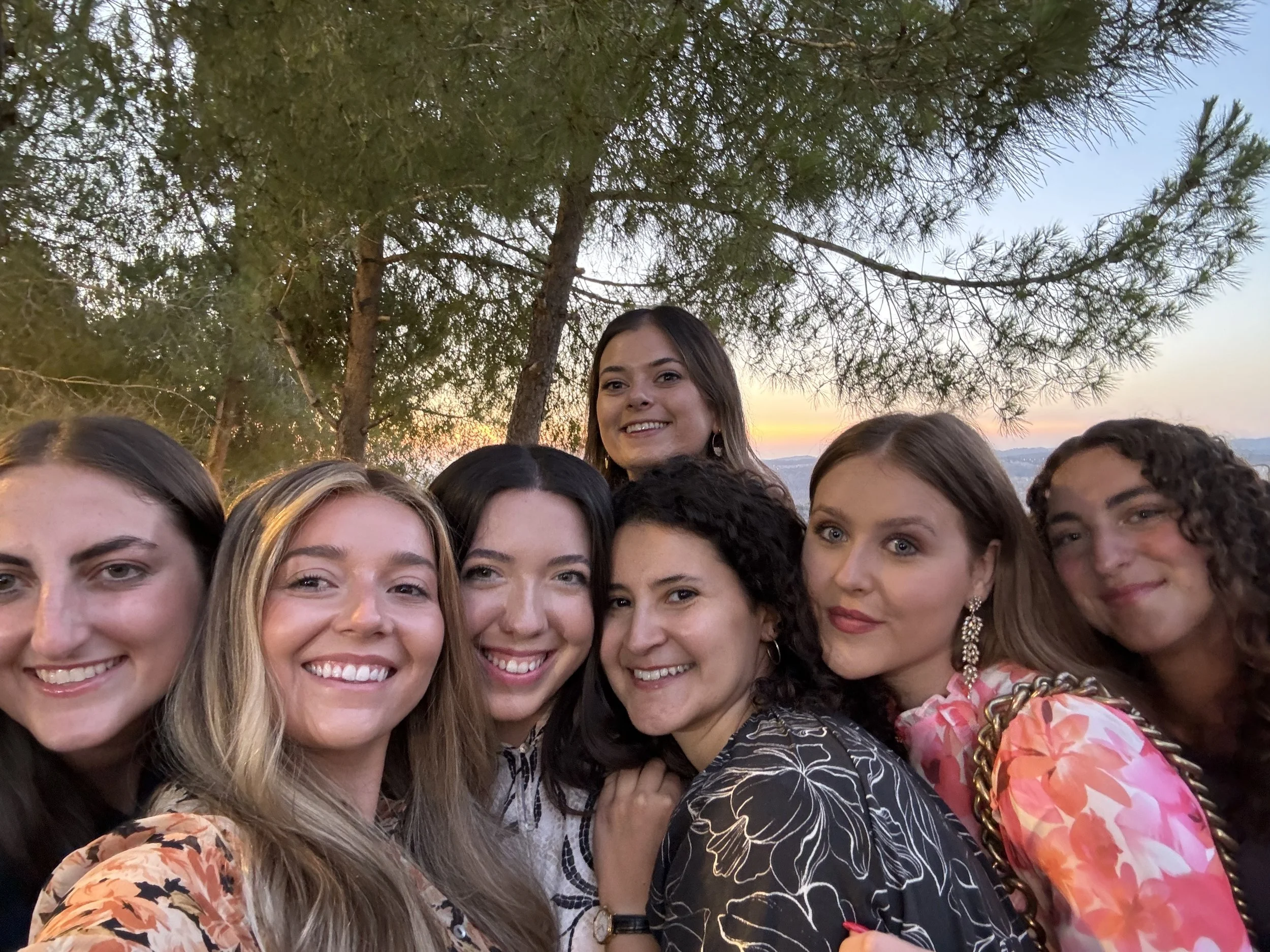 A group of women and a teenage girl smiling outdoors during sunset, with a background of trees and mountain ranges.