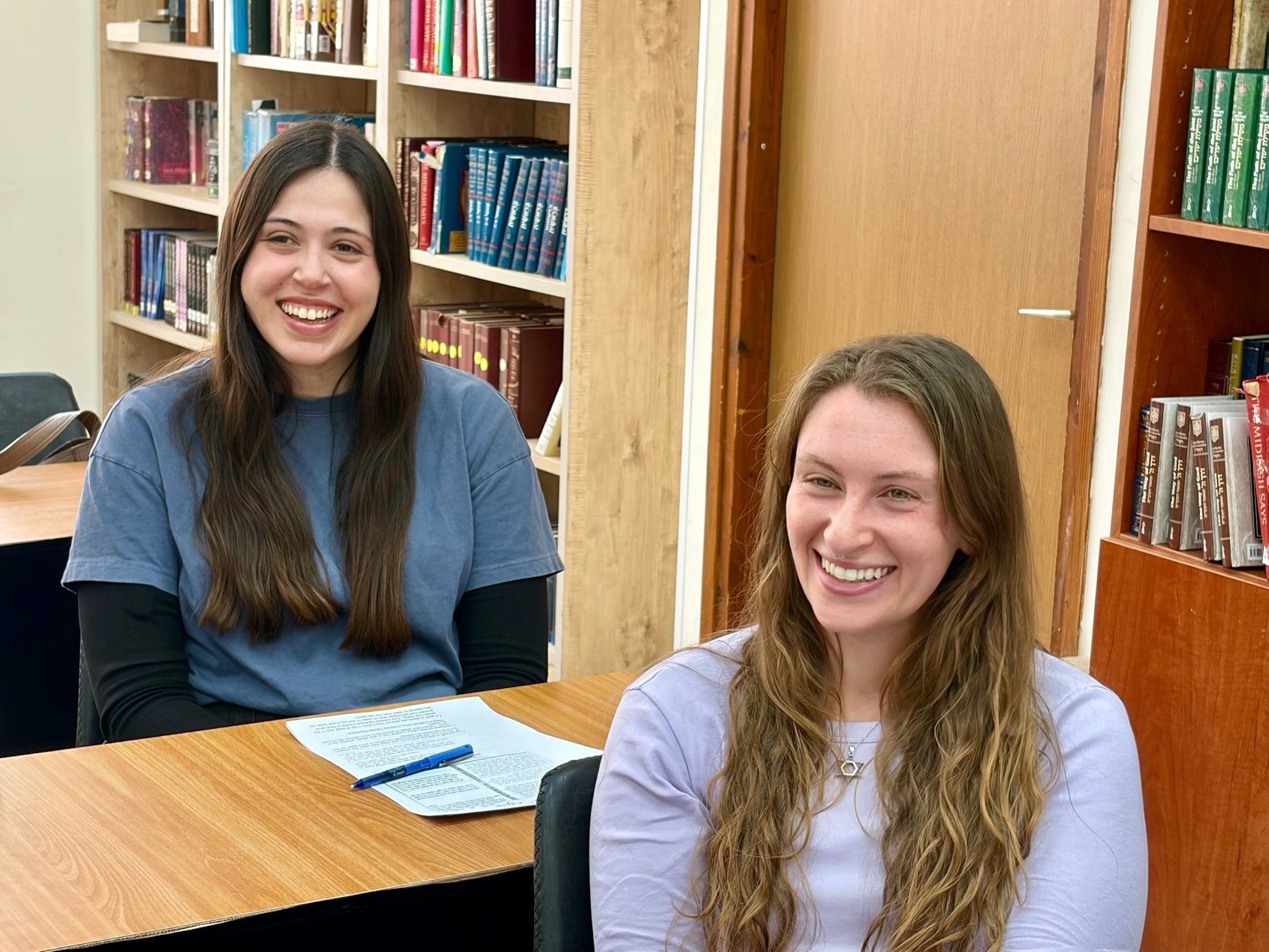 Two young women sitting at a wooden table in a library or classroom, smiling at the camera. Bookshelves filled with books are in the background. One woman has dark hair and a blue shirt, and the other has light brown hair and a lavender shirt. There are papers and pens on the table.