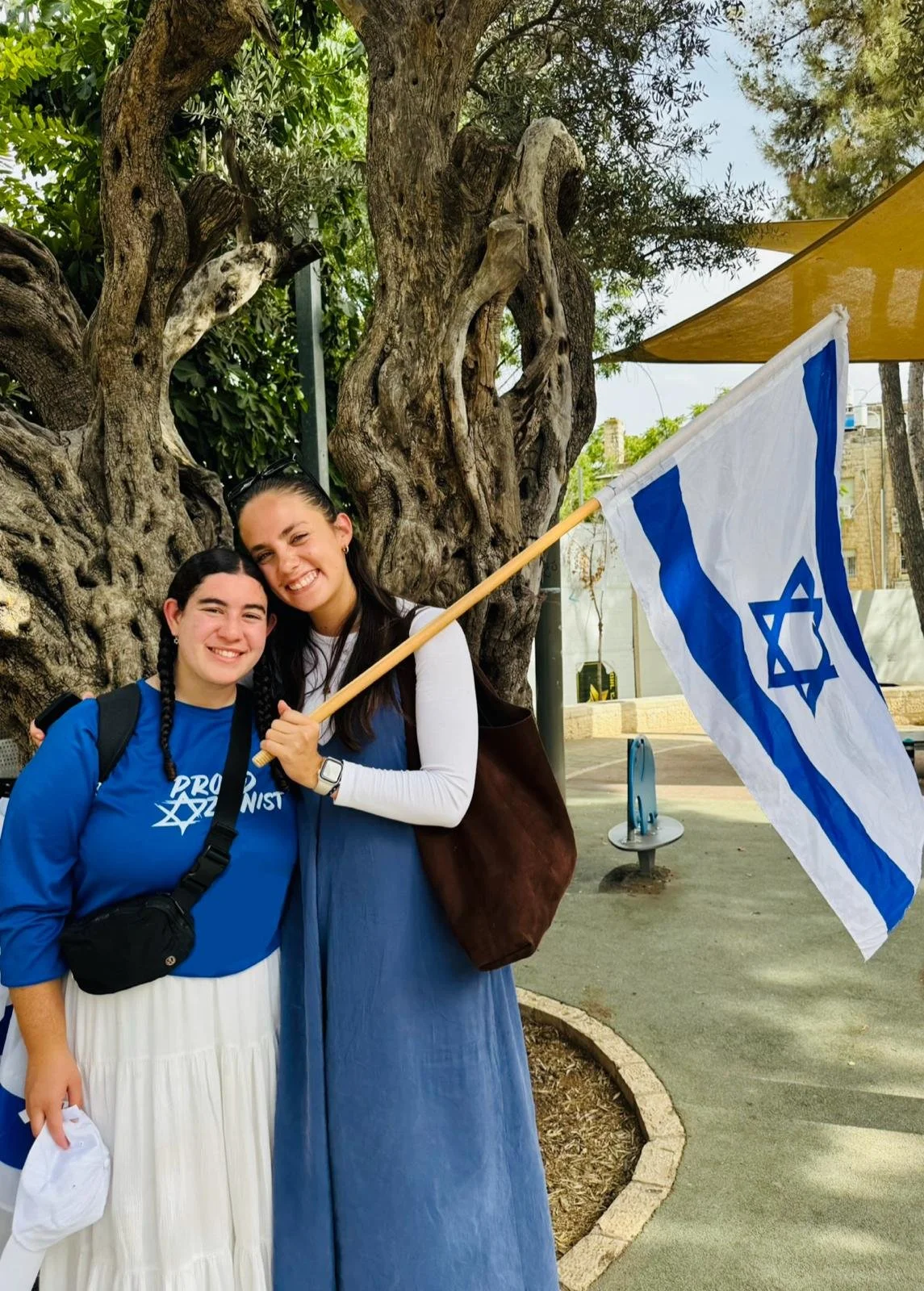 Two women smiling and holding an Israeli flag outdoors near a large gnarled tree. One woman wears a blue shirt with white text and a black bag, while the other wears a long blue dress with a white long sleeve shirt underneath. They are in a park or public space with benches and trees.