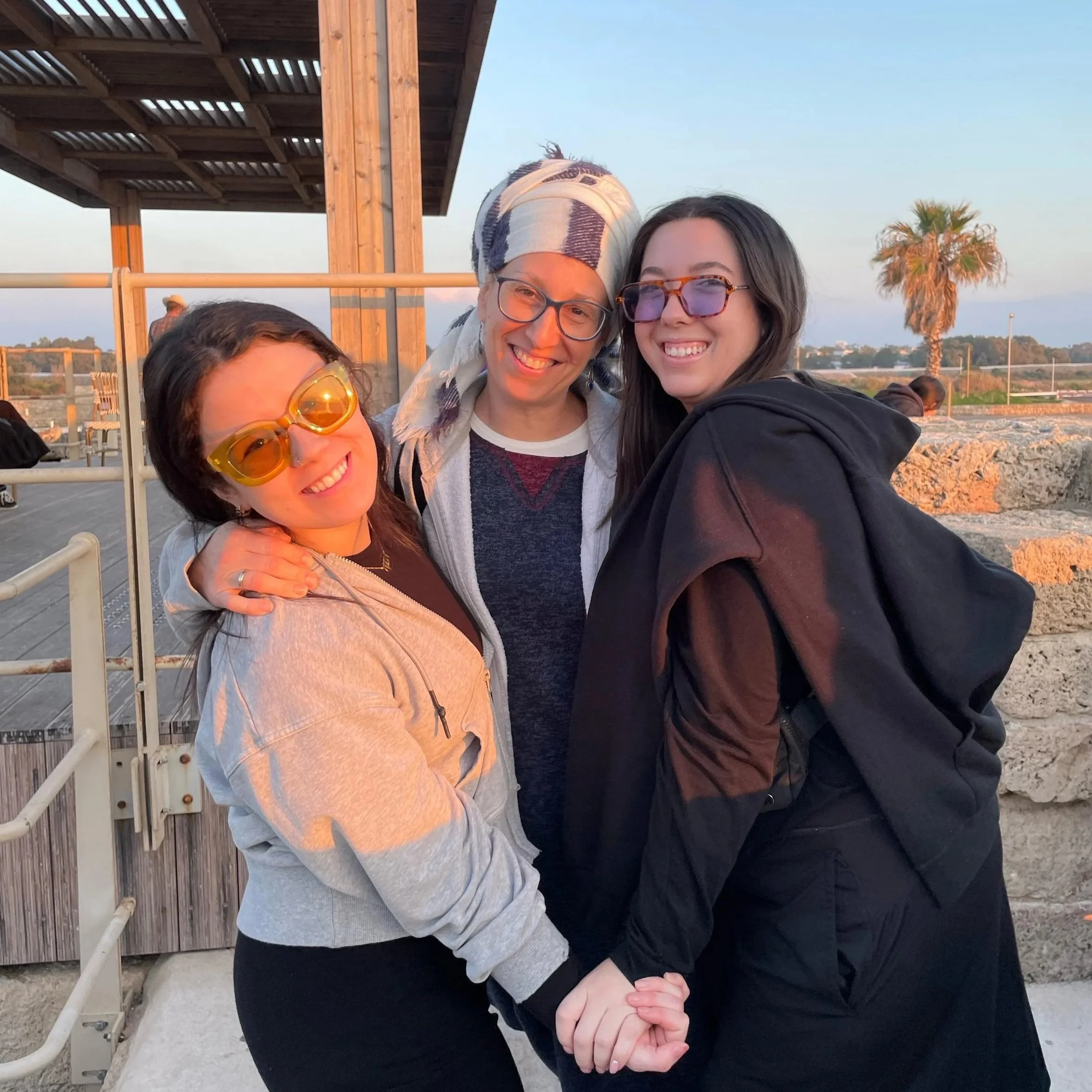 Four women smiling and holding hands together outdoors near a beach or waterfront with clear blue sky and palm trees in the background.