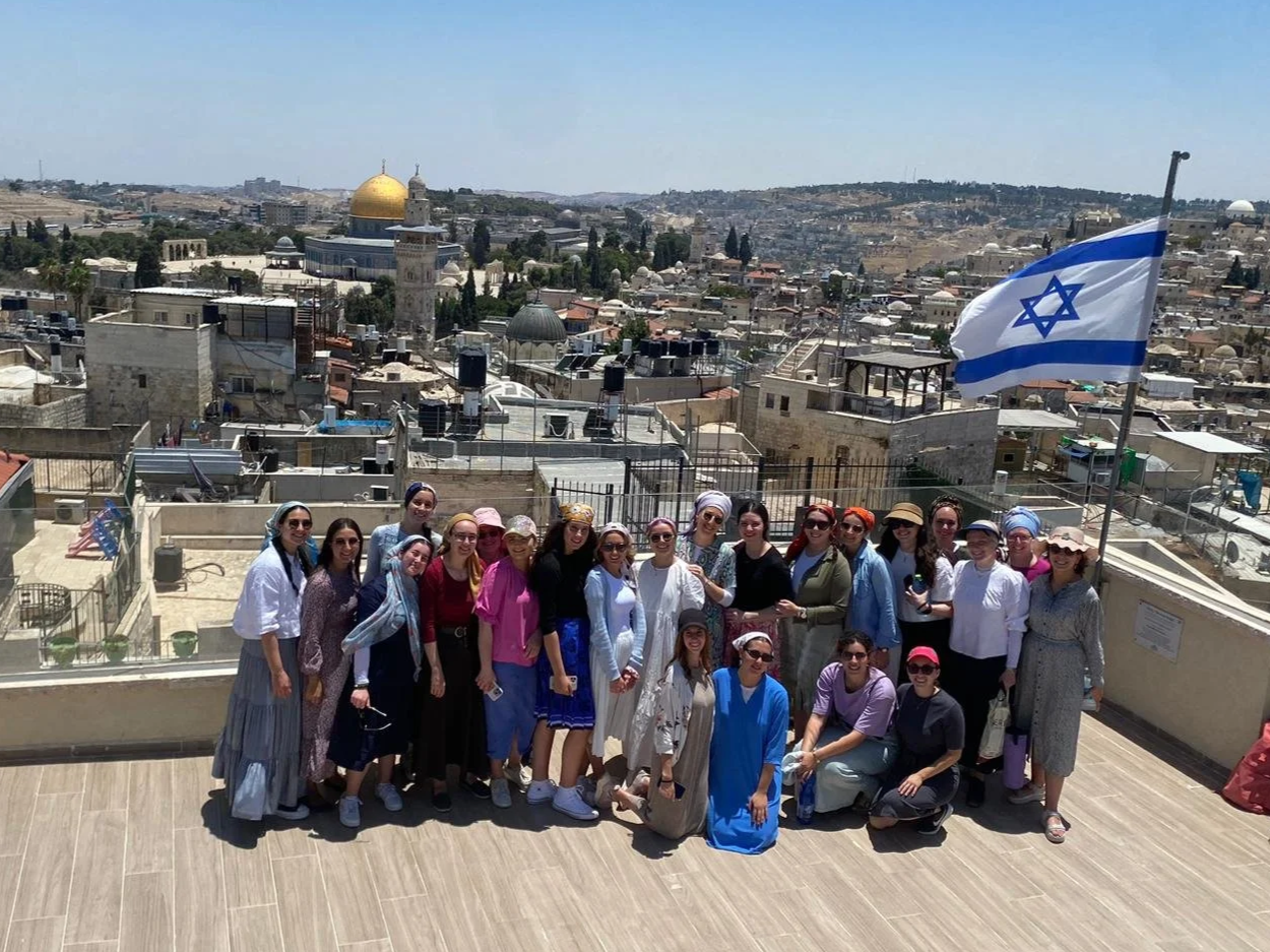 Group of women on a rooftop terrace with a view of Jerusalem's skyline, including the Dome of the Rock, and an Israeli flag.