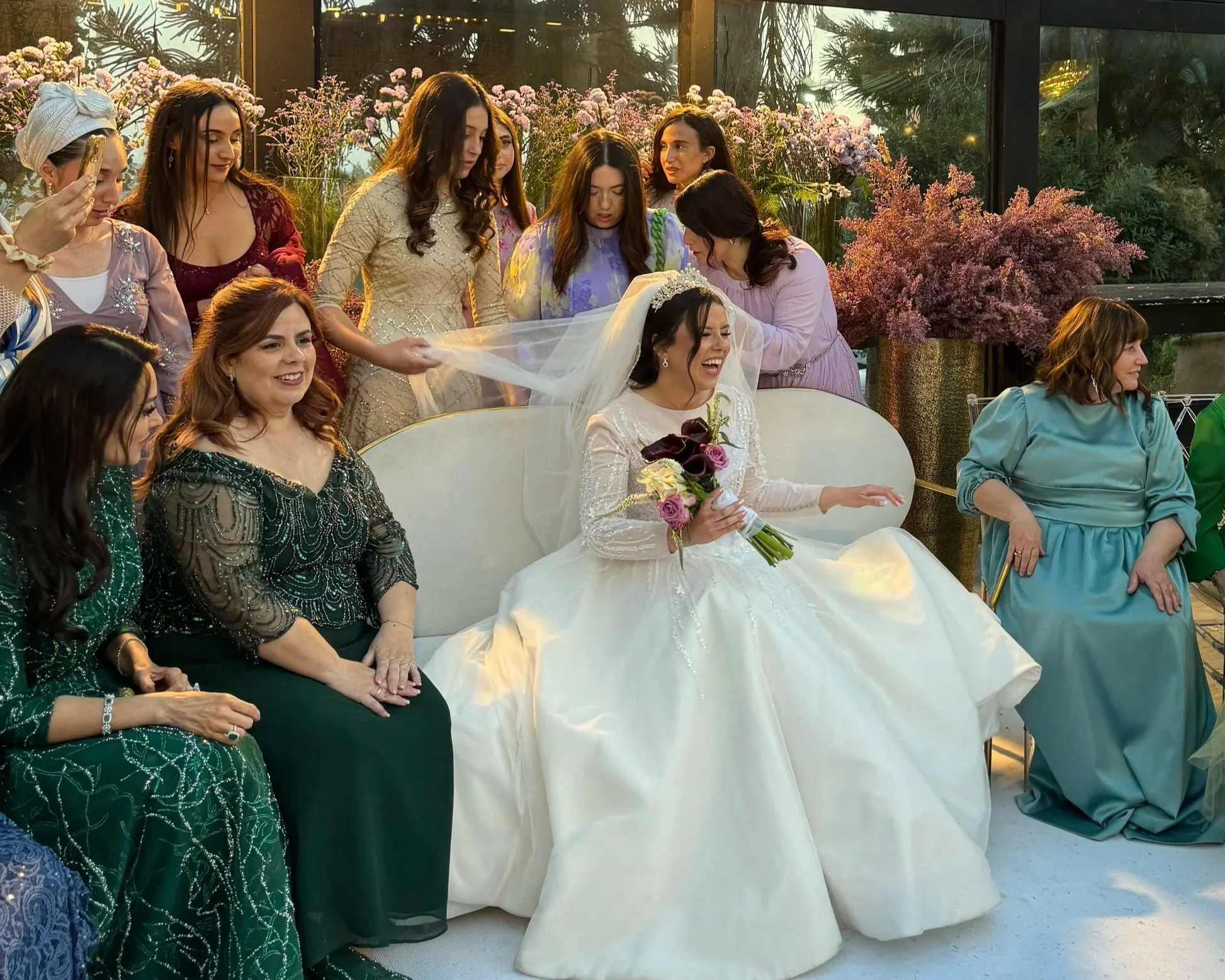 A group of women gathered around a bride on her wedding day. The bride is sitting smiling and holding a bouquet of flowers. The women are dressed in elegant, colorful dresses, with some touching or adjusting the bride's veil. The setting is decorated with pink flowers and greenery, with large glass windows in the background allowing natural light to fill the room.