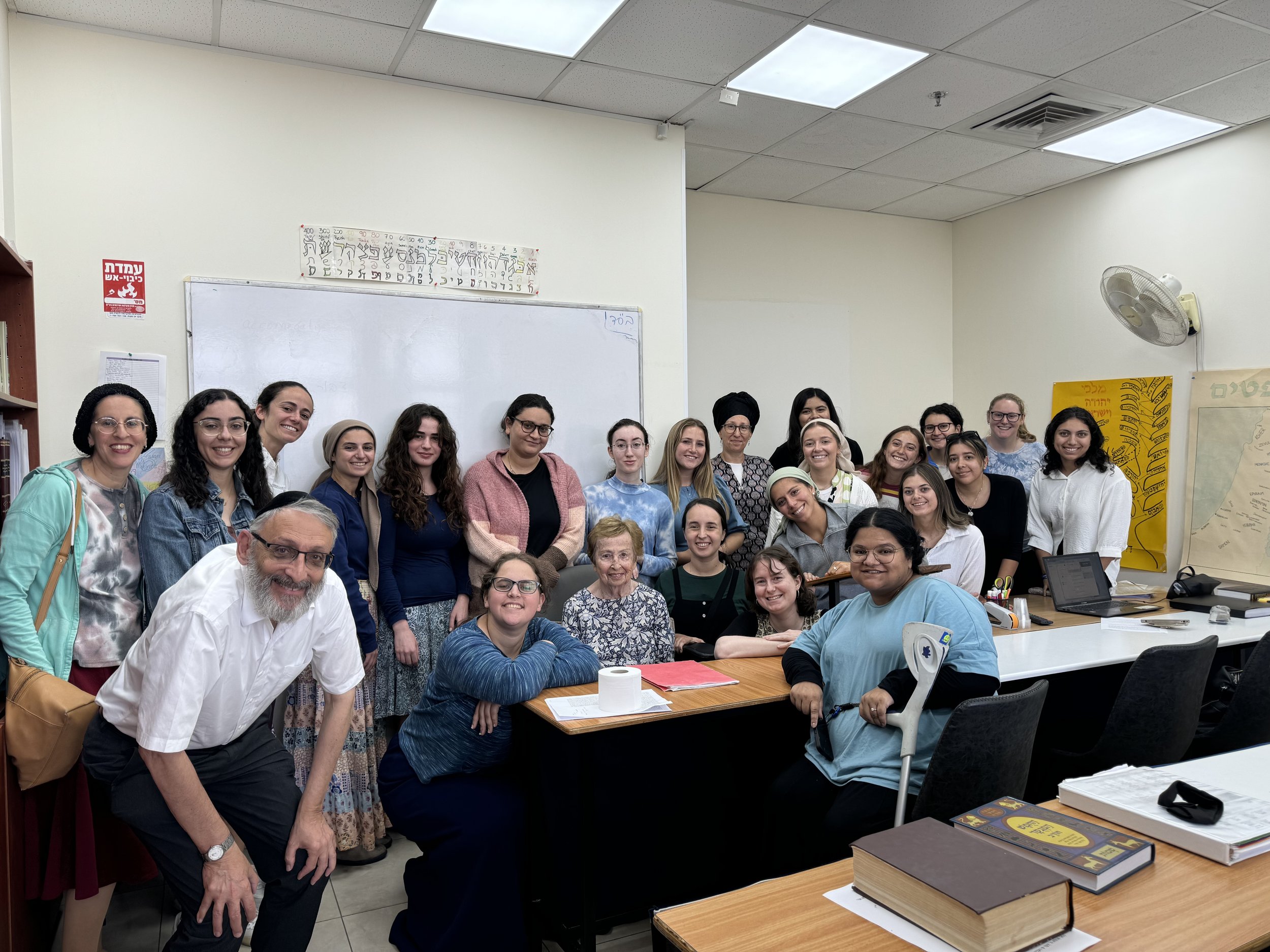 Group of diverse women and a man smiling and posing in a classroom with books, a whiteboard, and educational posters.
