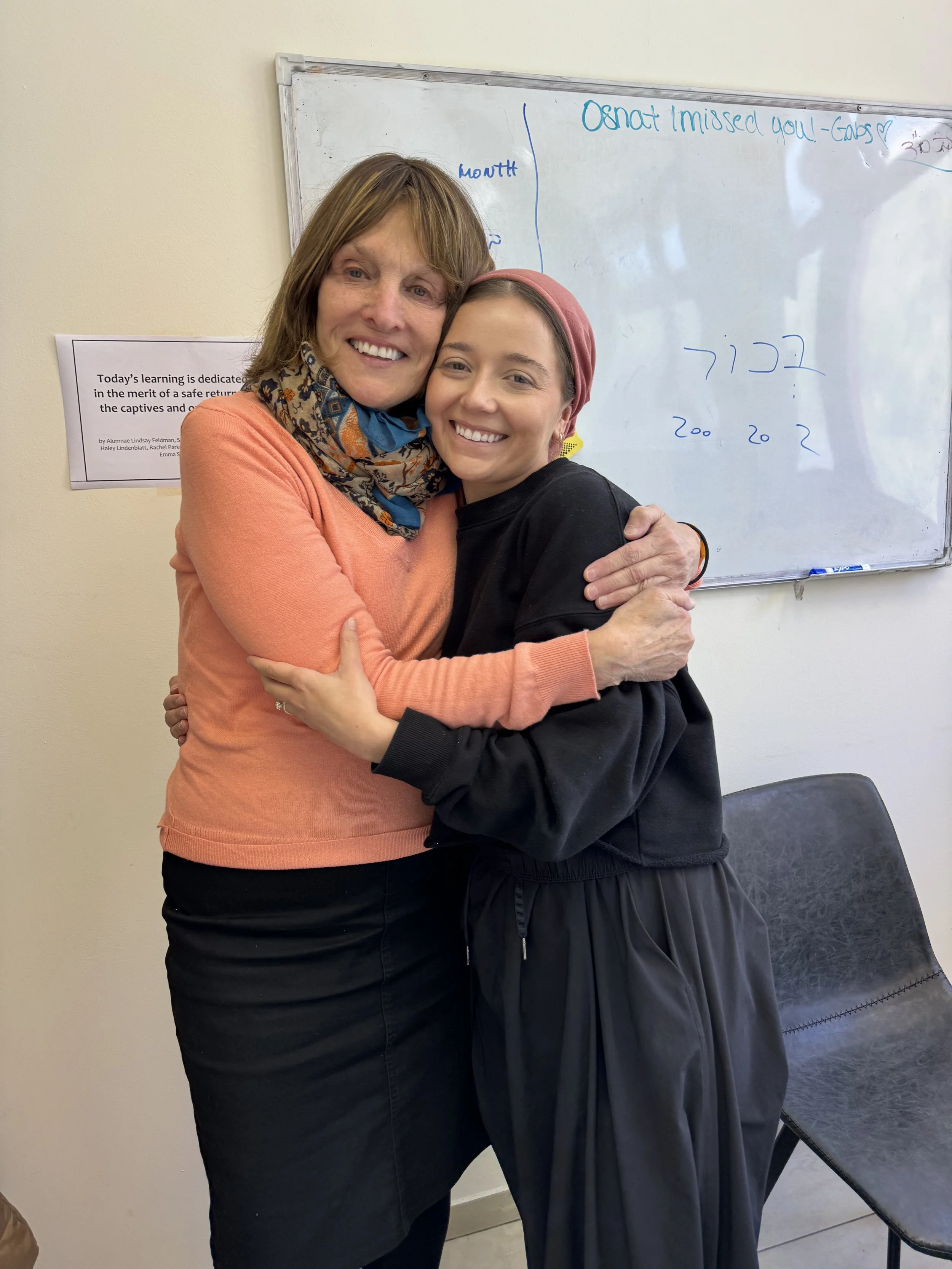 Two women hugging in a classroom, smiling at the camera. One is wearing an orange sweater and a colorful scarf, the other is wearing a black jacket and red headscarf. A whiteboard with handwritten notes is in the background.