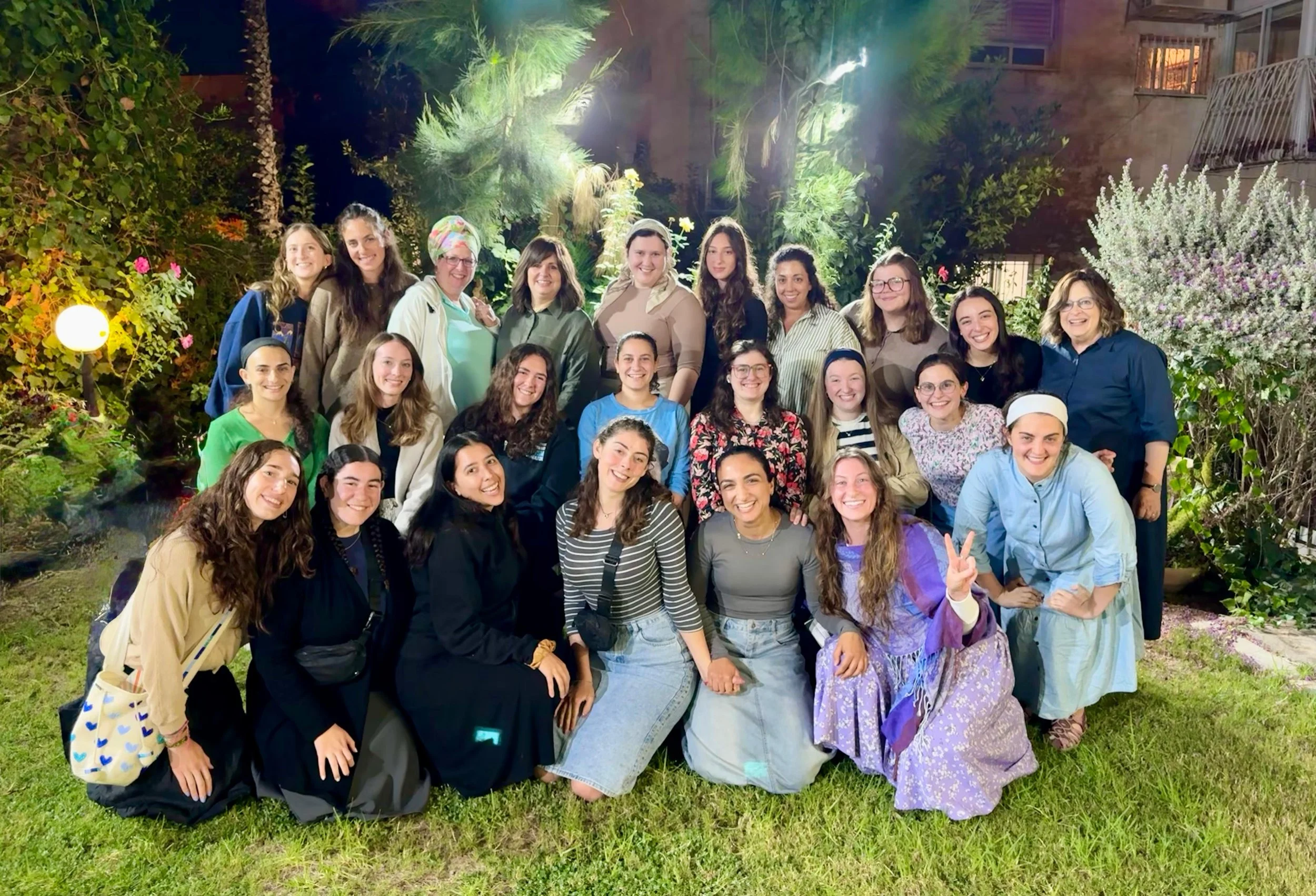 Group of women and girls posing together outdoors at night, standing on grass with trees and plants in the background.