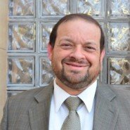 Portrait of a man with short dark hair, beard, wearing a gray suit and light-colored tie, smiling at the camera, standing in front of a glass block wall.