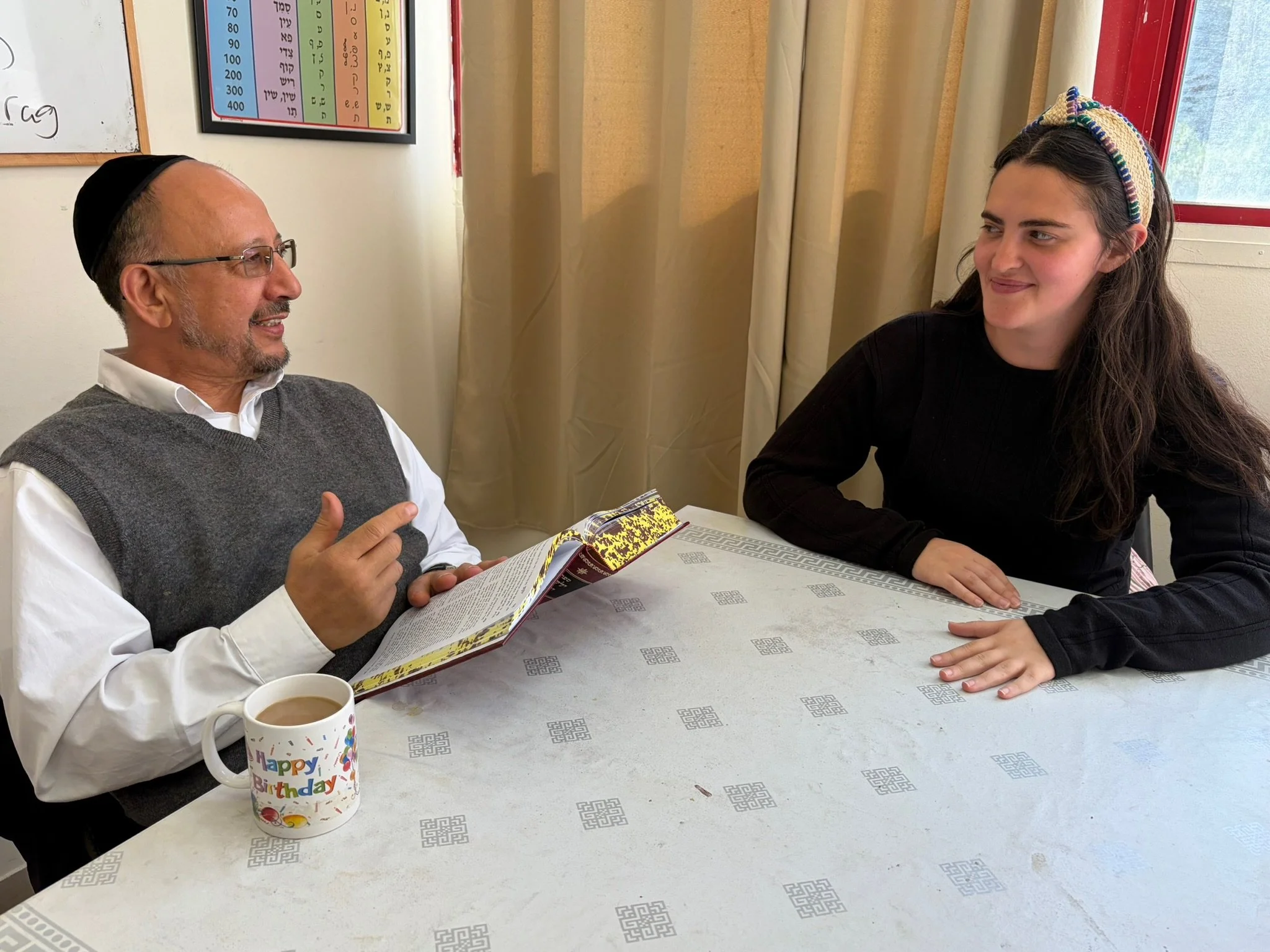 A man wearing glasses and a kippah sits at a table with a young woman who has long brown hair and a colorful headband. They appear to be having a conversation. There is a birthday mug and a cup of coffee on the table, and a colorful chart with Hebrew letters and numbers on the wall behind them.