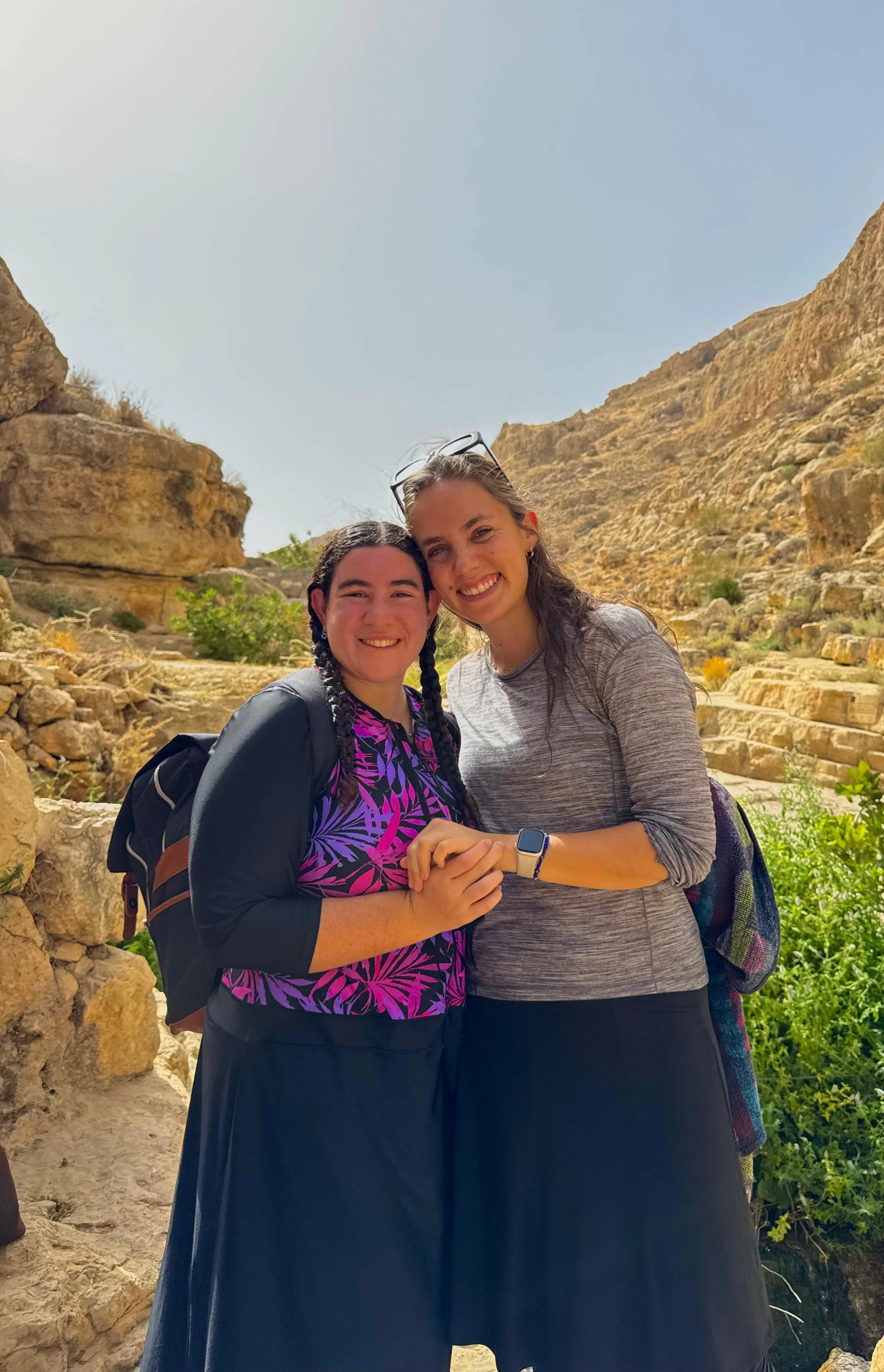 Two women smiling and holding hands, outdoors in a rocky, mountainous landscape with greenery and a clear sky.