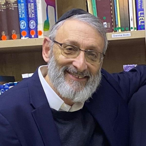 Smiling older man with glasses and a gray beard, wearing a dark blazer and white shirt, in a library or bookstore with shelves of books in the background.