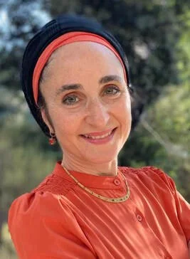 A woman smiling outdoors, wearing a red blouse, a headband, and jewelry.