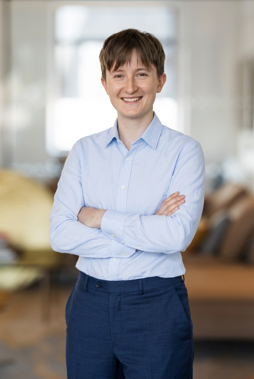 Smiling young woman in light blue button-up shirt and dark blue pants standing with arms crossed in an office environment.