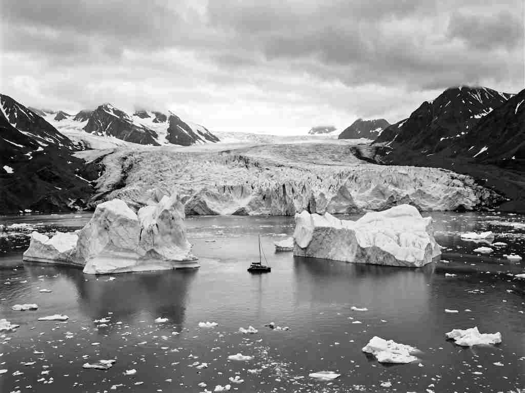 Black and white photo of a glacier calving into a body of water with icebergs and mountains in the background.