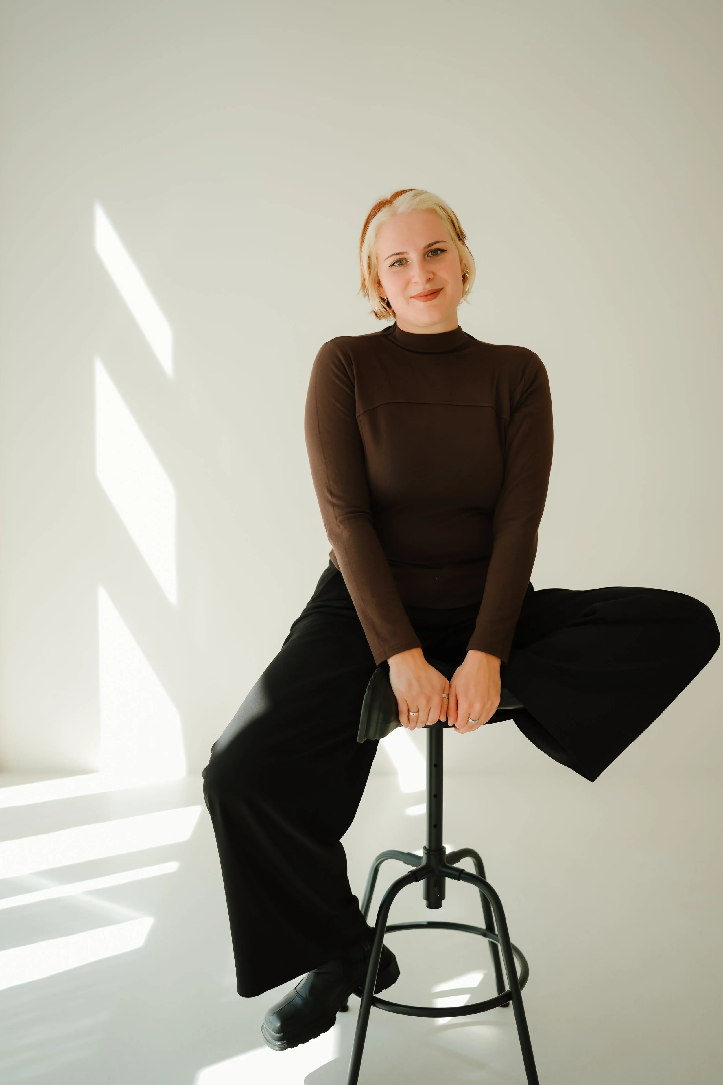 Porträt einer jungen Frau mit blondem, kurz geschnittenem Haar, sitzend auf einem Hocker vor weißem Hintergrund, trägt einen schwarzen Turtleneck und schwarze Hosen, lächelt leicht.