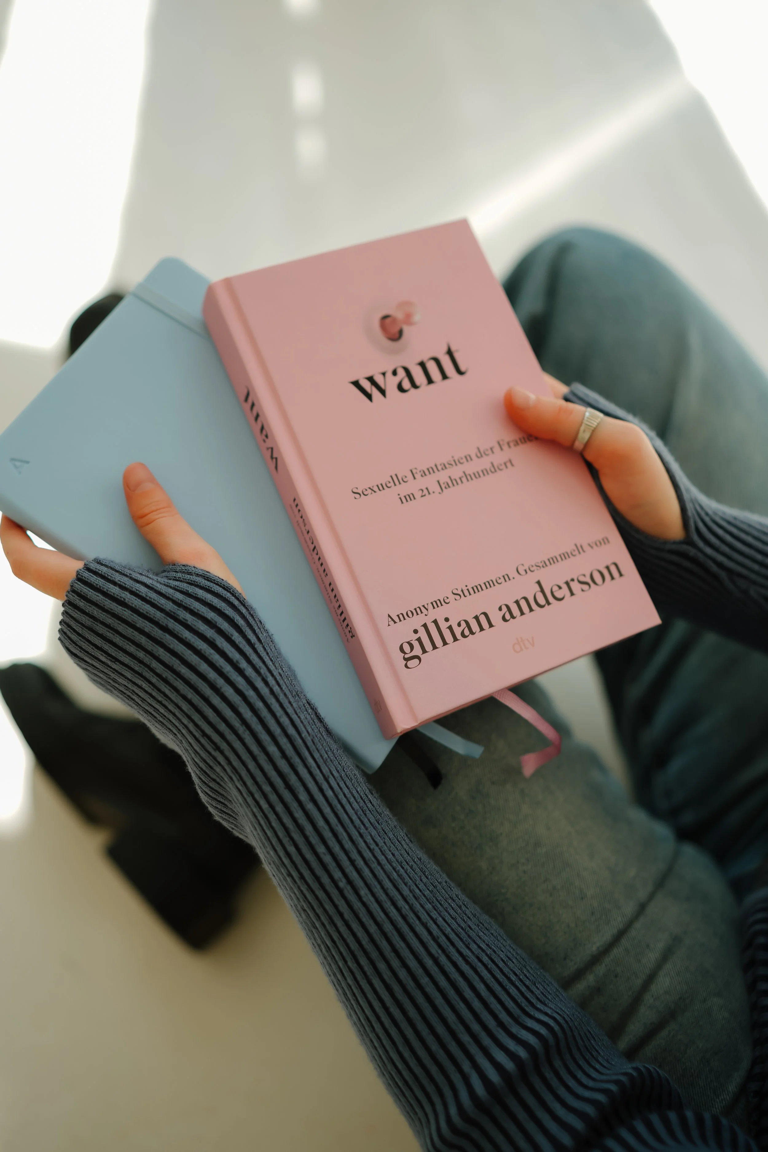 Hand hält zwei Bücher, eines rosa mit dem Titel "want" und eine andere blaue, beim Durchsehen eines Wartebereichs, vermutlich in einer Zugsstation.