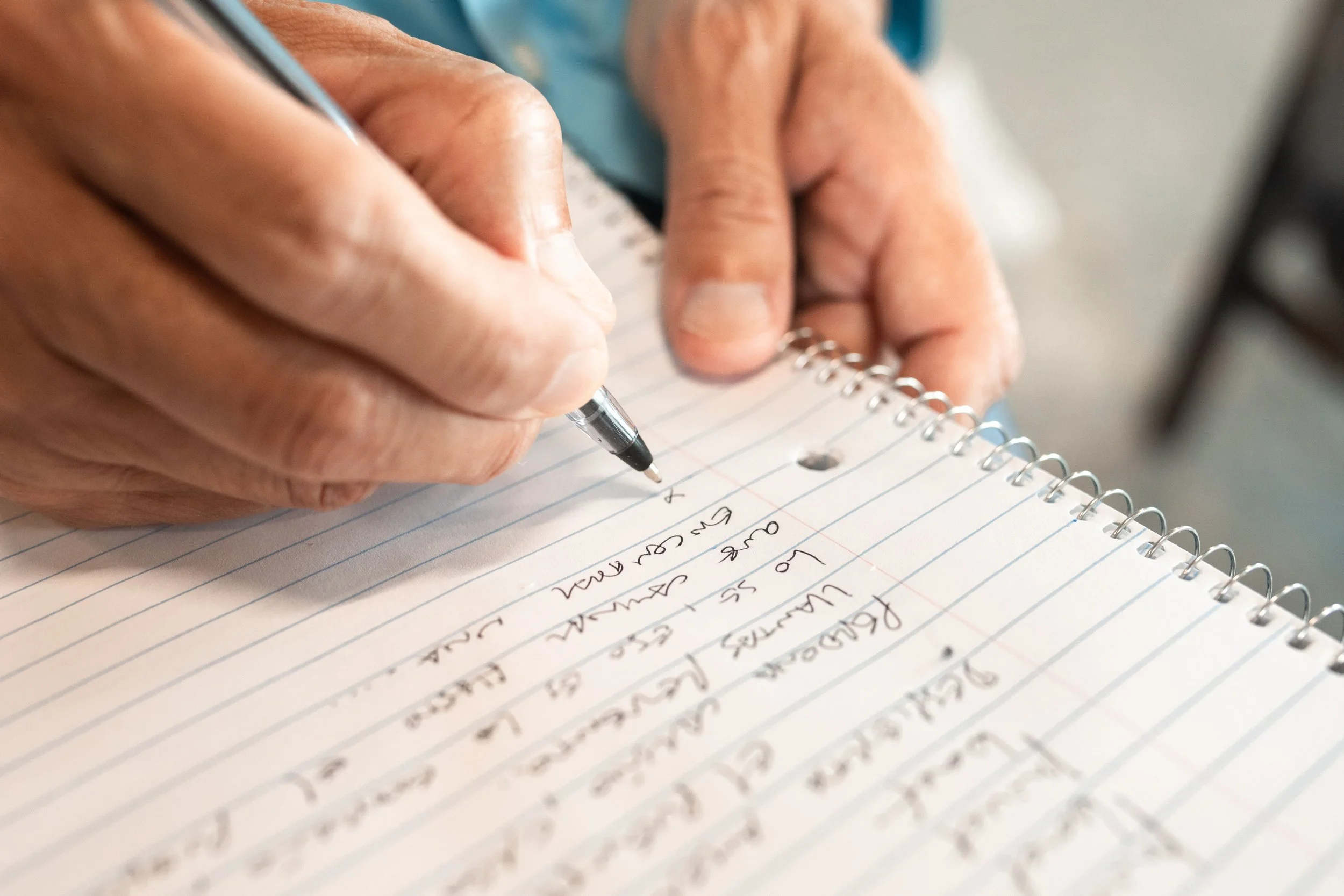 Close-up of a person writing notes in a spiral notebook with a black pen, with handwritten text visible on lined paper.