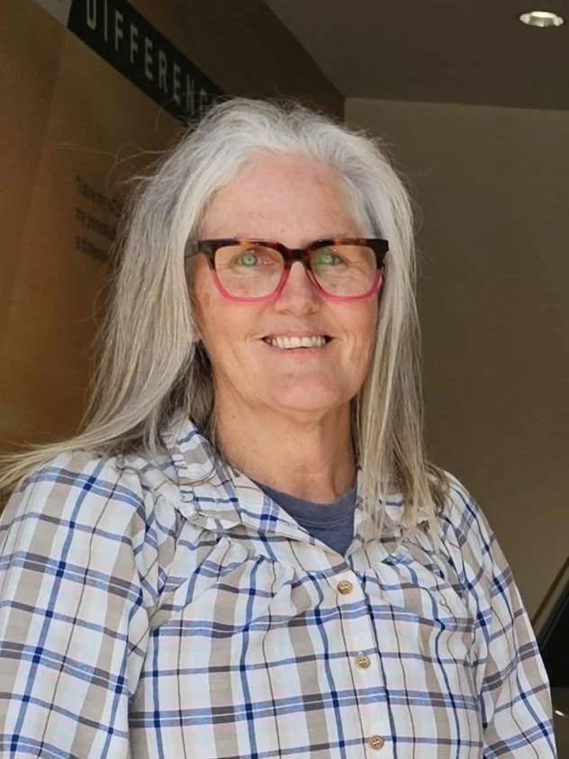 Geri Bennett, a woman with long grey hair, wearing glasses with colourful frames, a plaid shirt, and standing indoors.