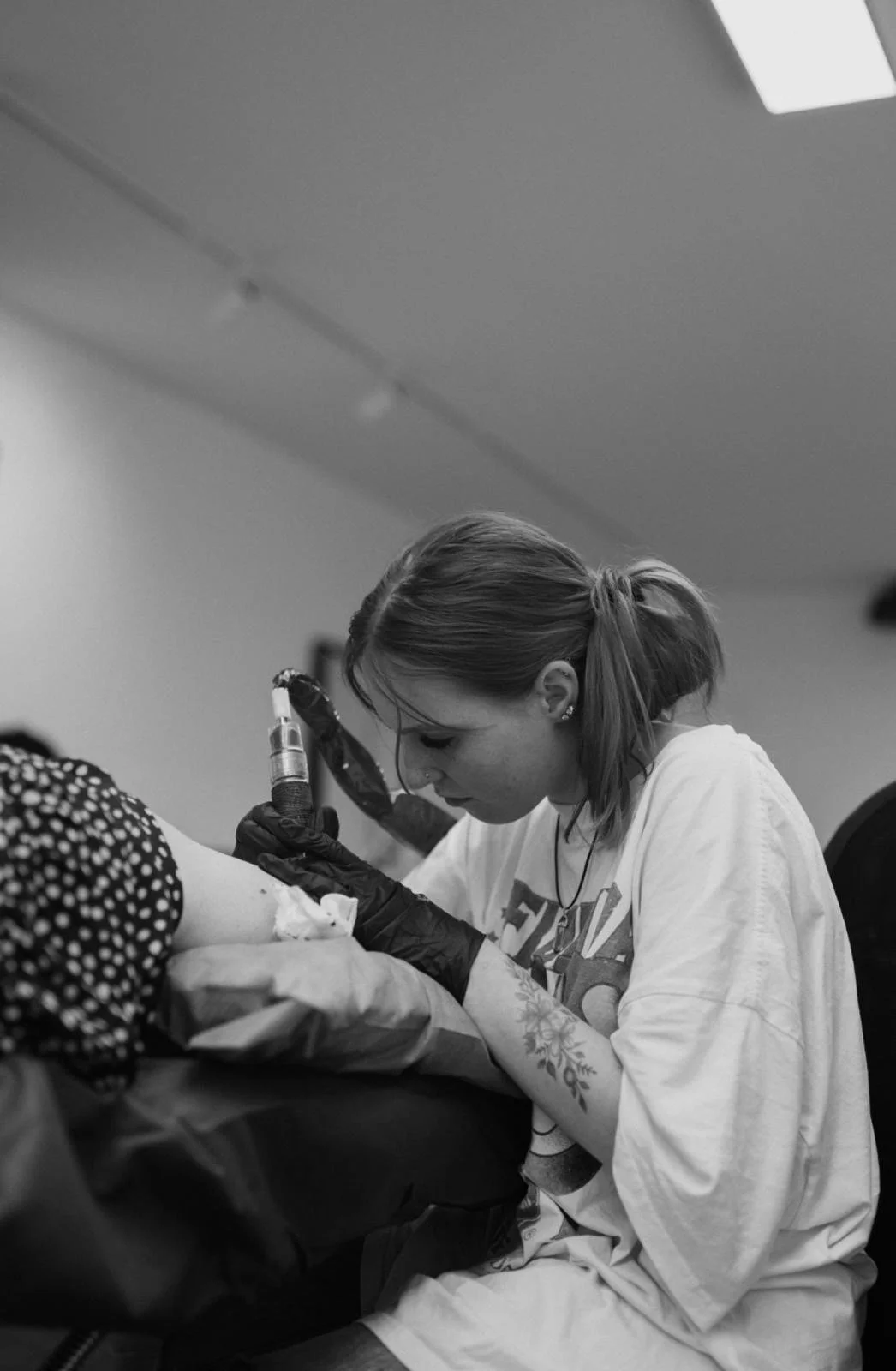A woman with tattoos on her arm, wearing a casual T-shirt, is tattooing a person's arm in a tattoo studio. She is focused on her work, wearing gloves, holding a tattoo machine, and leaning over the person's arm, which is draped with a cloth.