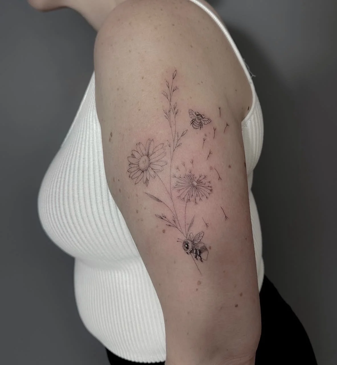 A tattoo of a floral design with a bee on a person's upper arm, showing delicate line art of flowers, a bee, and a small insect.