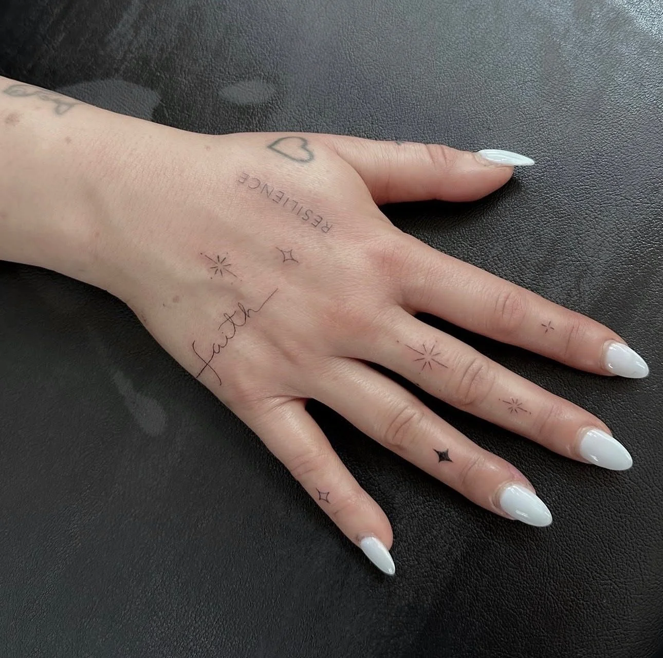 A hand with white painted nails resting on a black surface. The hand has various small tattoos, including the word 'faith,' a heart, stars, and the word 'RESTIENCE'.