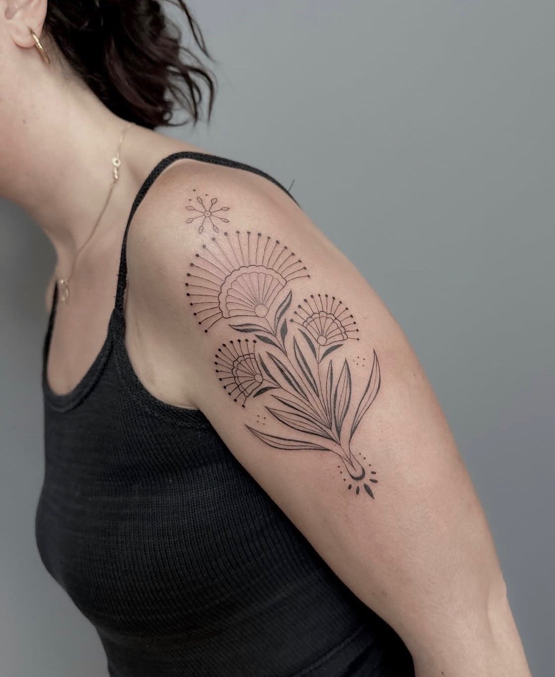 A person's upper arm and shoulder featuring a black line art floral tattoo with leaves and flowers in a minimalist style.