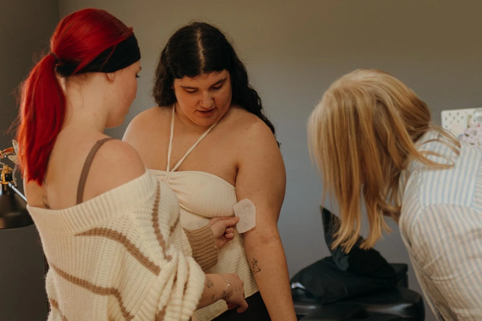 A woman with dark hair is having a tattoo applied to her upper arm by a person with red hair. The woman is wearing a cream-colored off-the-shoulder top and appears to be in a tattoo parlor, with another woman with blonde hair observing.