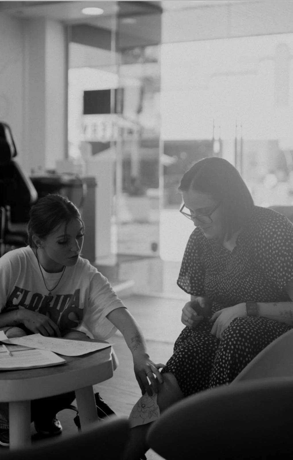 A woman in glasses and a polka dot dress sits in a chair, getting a tattoo on her foot by a young woman with a tattoo and wearing a Florida shirt, inside a tattoo studio.