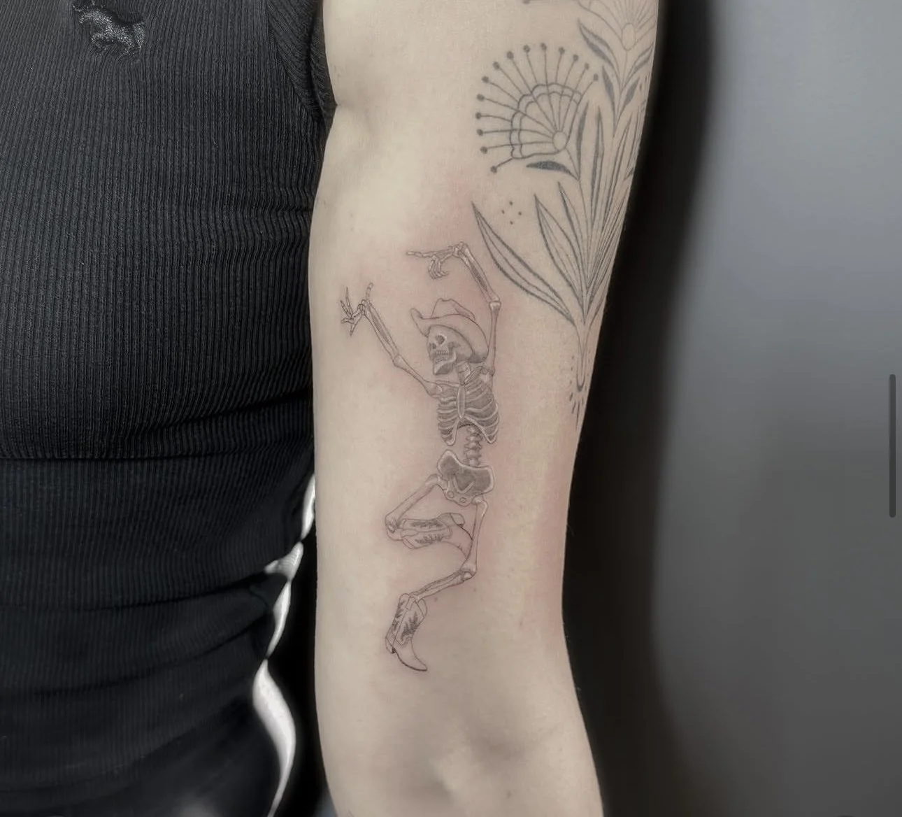 A tattoo of a dancing skeleton wearing a cowboy hat on a person's upper arm, along with a large outline of a plant or floral design on the same arm.
