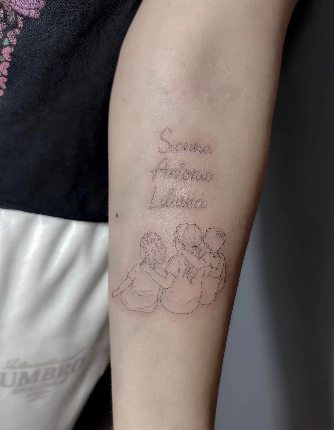 Tattoo on person's arm featuring the names Sienna, Antonio, and Liliana in cursive, with a line drawing of three children sitting together, one with curly hair, one with long hair, and one with short hair, arms around each other.