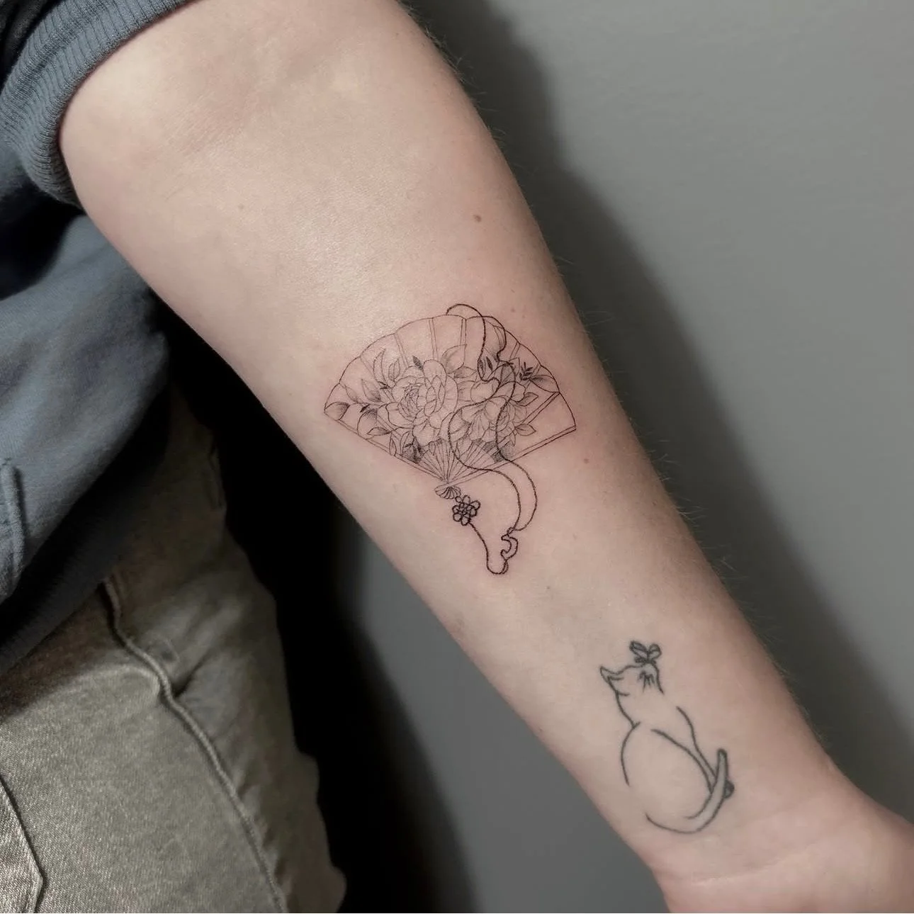 Tattoo of a fan with flowers inside on a person's forearm, with a simple line drawing of a cat holding a flower below it.