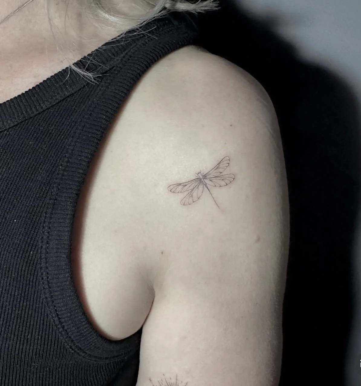 A close-up of a person's shoulder and upper arm, showing a delicate dragonfly tattoo on their shoulder, with the person wearing a dark sleeveless top.