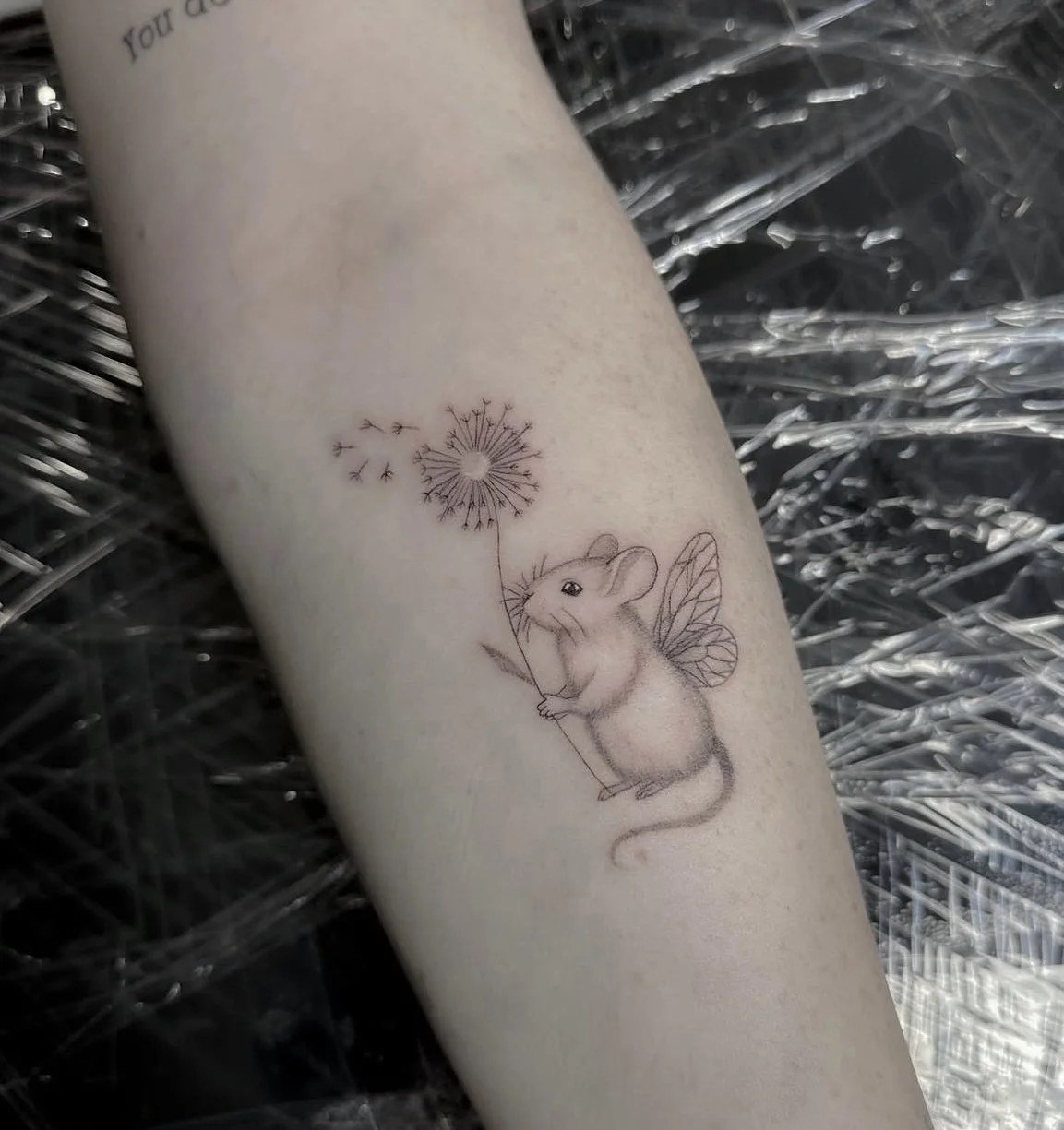 Tattoo of a mouse with fairy wings holding a dandelion, with a few seeds blowing away.