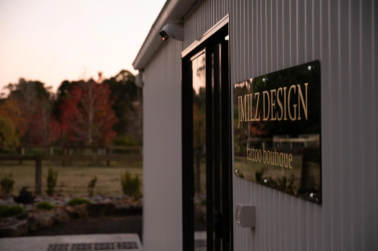 Close-up of a black sign on the side of a building labeled 'MJZ DESIGN tattoo boutique' with a background of trees and a sunset sky.
